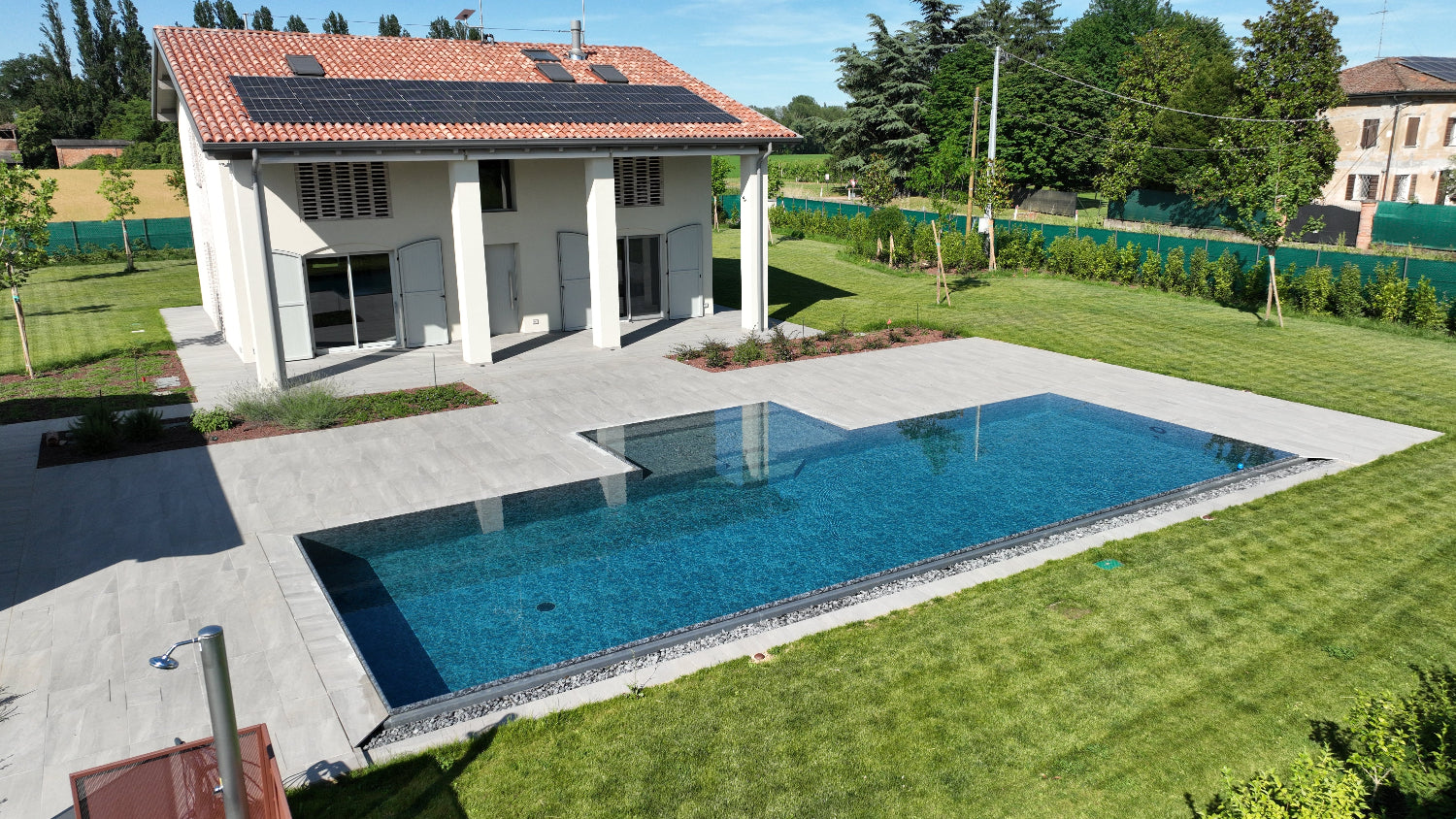 Piscina a Sfioro senza vasca di compenso, lato con muro a cascata e vaschette con sassi decorativi, effetto orizzonte. Rivestimento nero marmorizzato e spiaggia centrale.