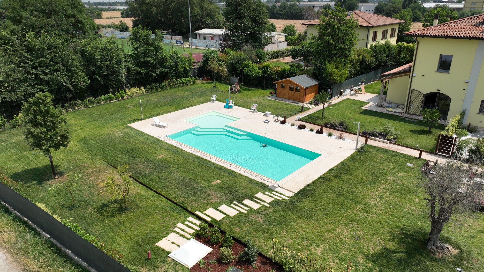 Piscina a forma libera con spiaggia angolare esterna e rivestimento sabbia, vaschette con sassi decorativi su lato lungo per effetto cascata