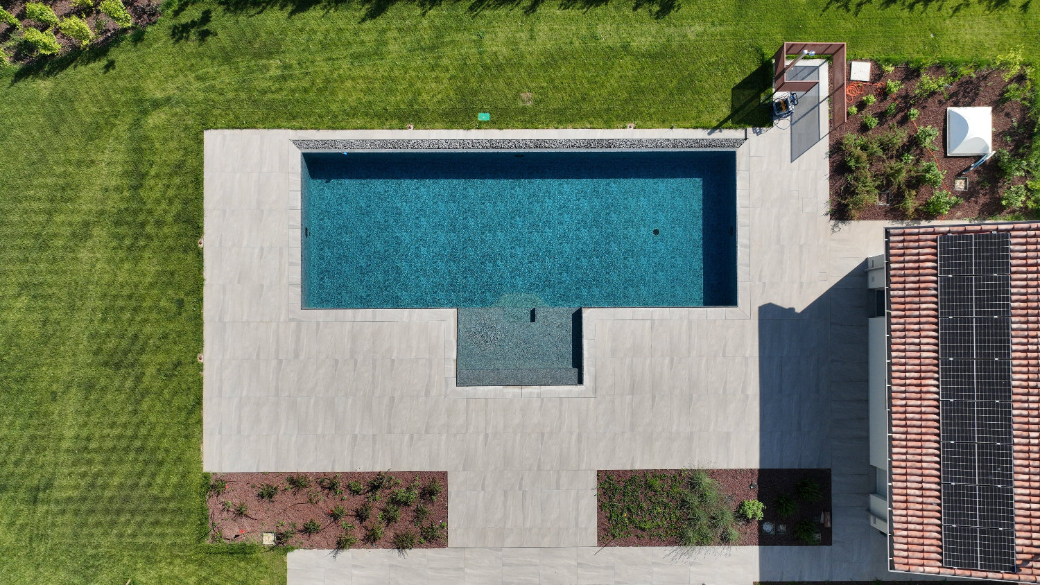 Piscina a sfioro con spiaggia esterna centrale, lato lungo con vaschette di sassi decorativi, rivestimento marmorizzato e pavimento grigio chiaro, riprese drone 