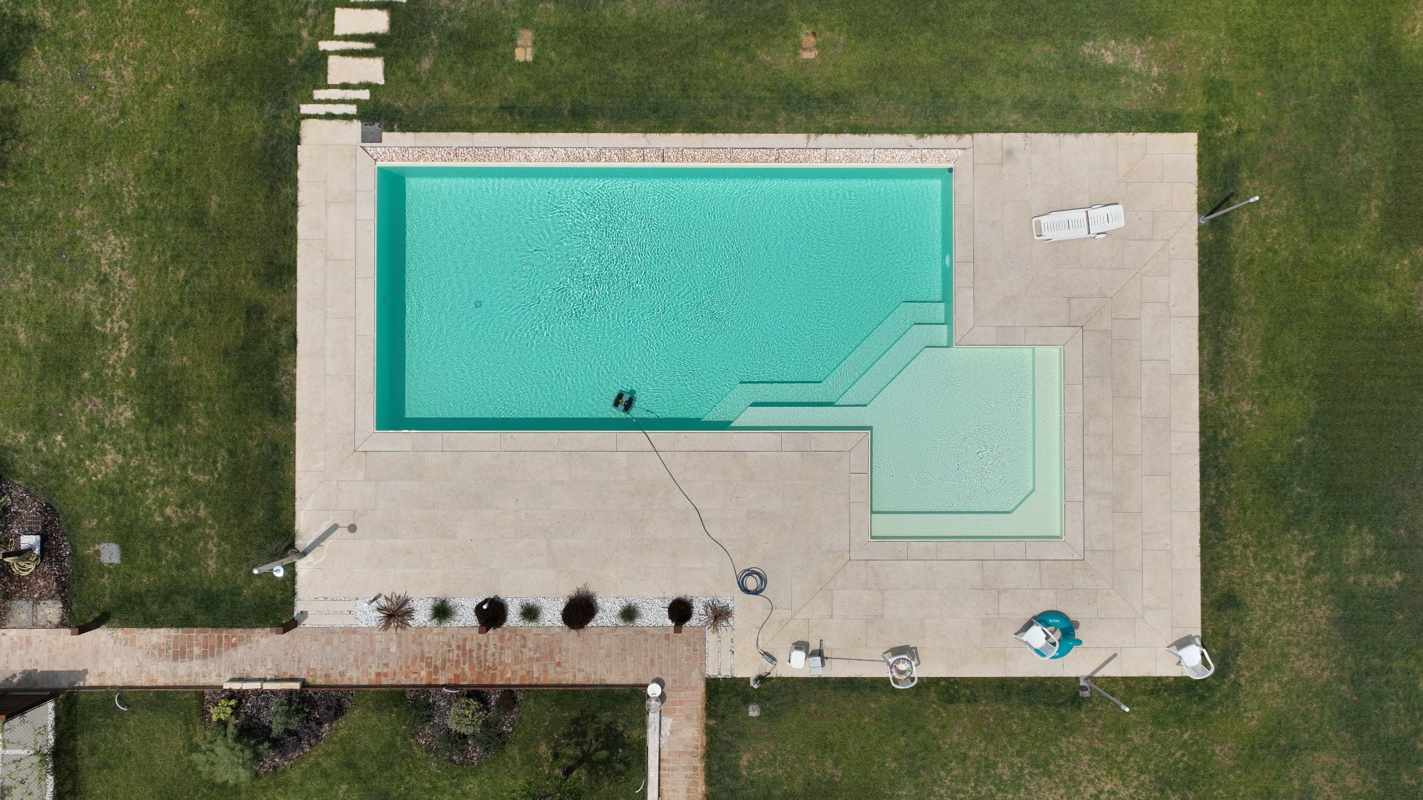 Piscina a sfioro dal design unico e moderno, rettangolare con bagnasciuga esterno in angolo e gradoni obliqui, vaschette con sassi decorativi su un lato lungo