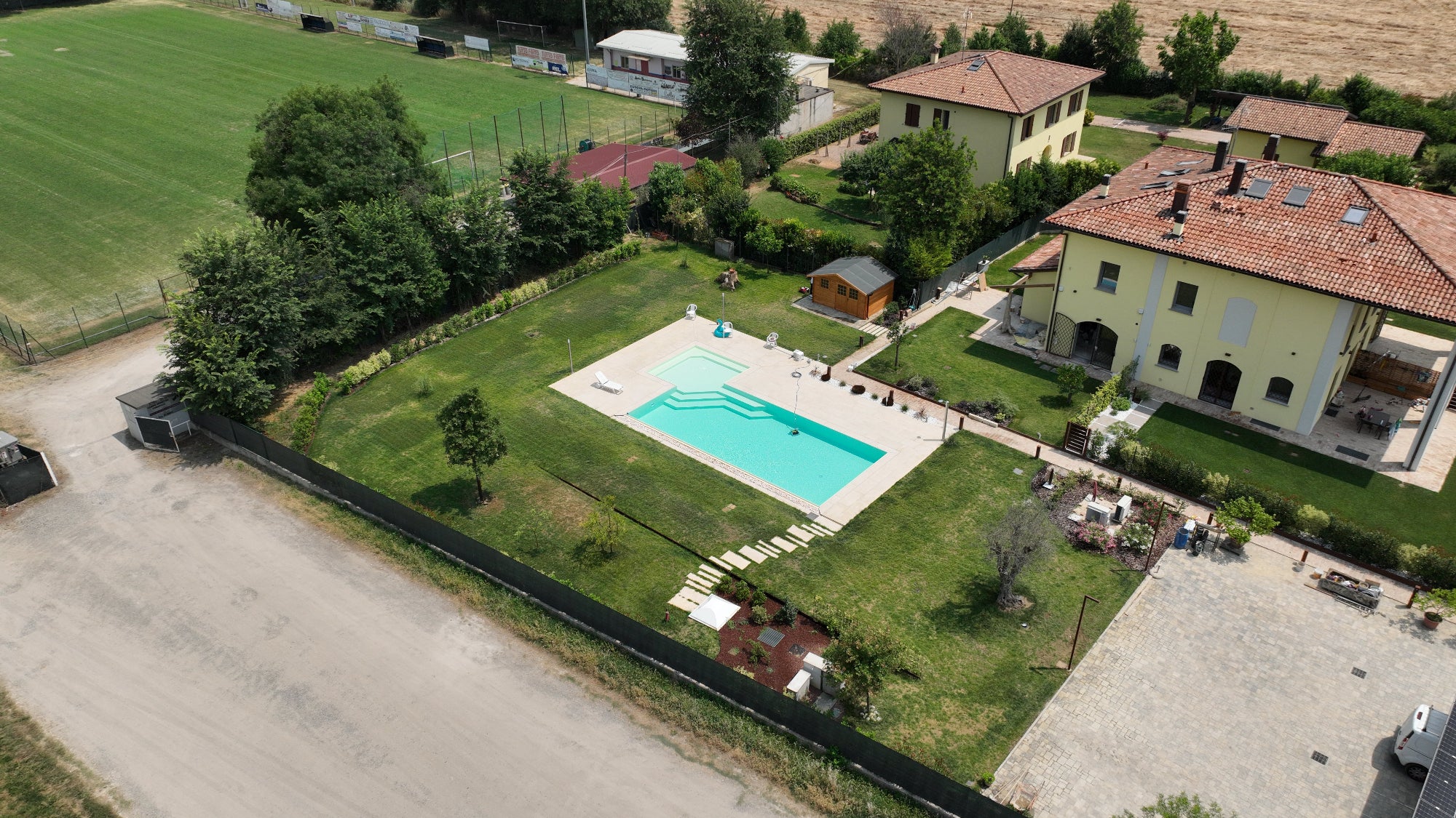 Piscina a sfioro senza vasca di compenso con vaschette in acciaio e sassi decorativi sul lato lungo, rivestimento color sabbia e zona relax angolare esterna in zona Bologna.