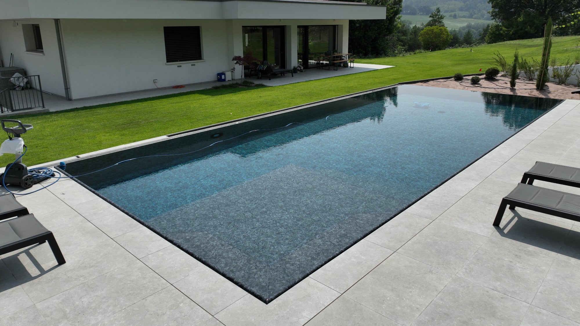 Piscina a sfioro senza vasca di compenso di esclusiva Zavatti, forma rettangolare con spiaggia interna, due lati con muro a cascata e vaschette con sassi decorativi effetto infinity, rivestimento nero marmorizzato