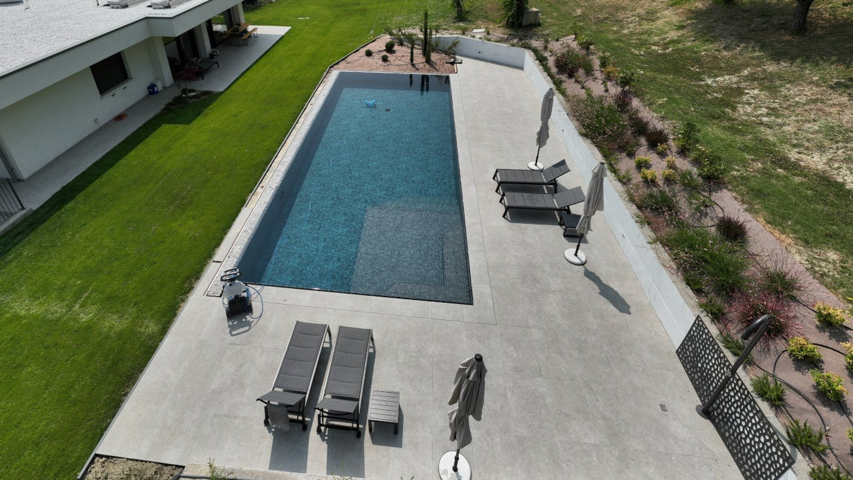 Piscina sfioro a Castellarano, dal design pulito e minimal, rivestimento nero marmorizzato in contrasto con pavimento chiaro e verde del prato. Vaschette con sassi su due lati per decoro moderno