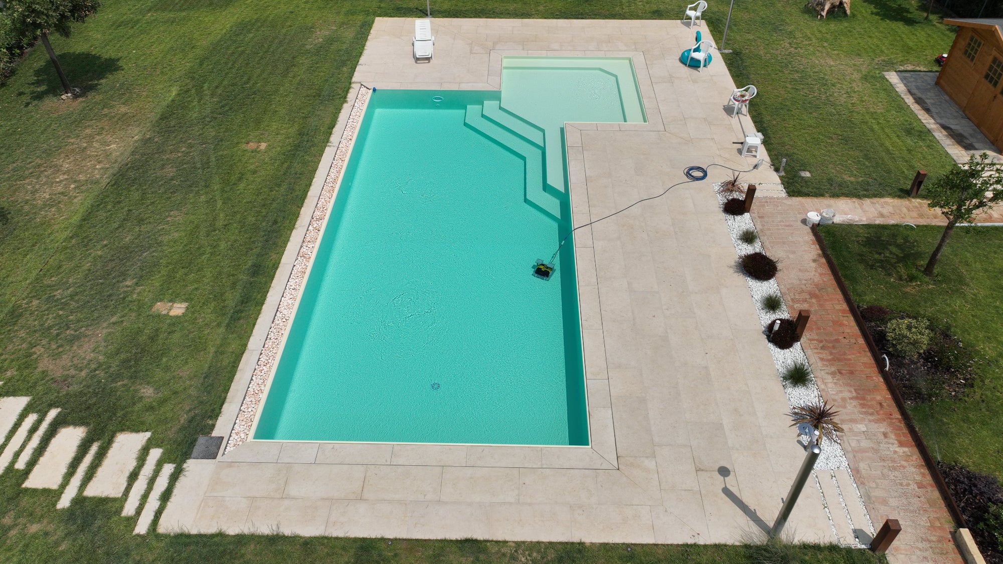 Piscina sfioro senza vasca di compenso immersa nel verde a Bologna, rivestimento color sabbia e vaschette decorative con sassi sul lato lungo 