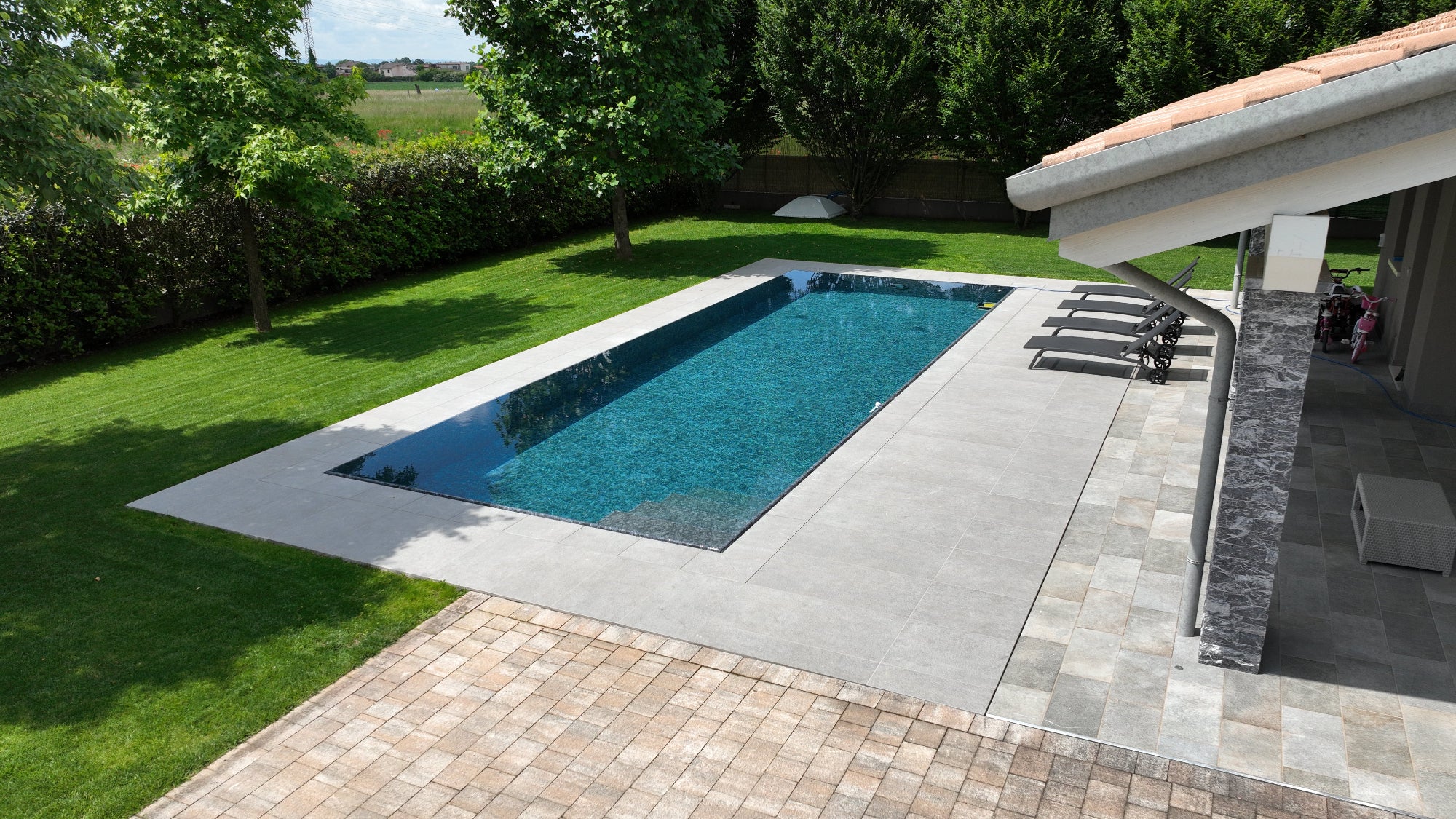 bellissima piscina a sfioro senza vasca di compenso con rivestimento nero effetto marmo, in contrasto con pavimento grigio chiaro e prato verde. 