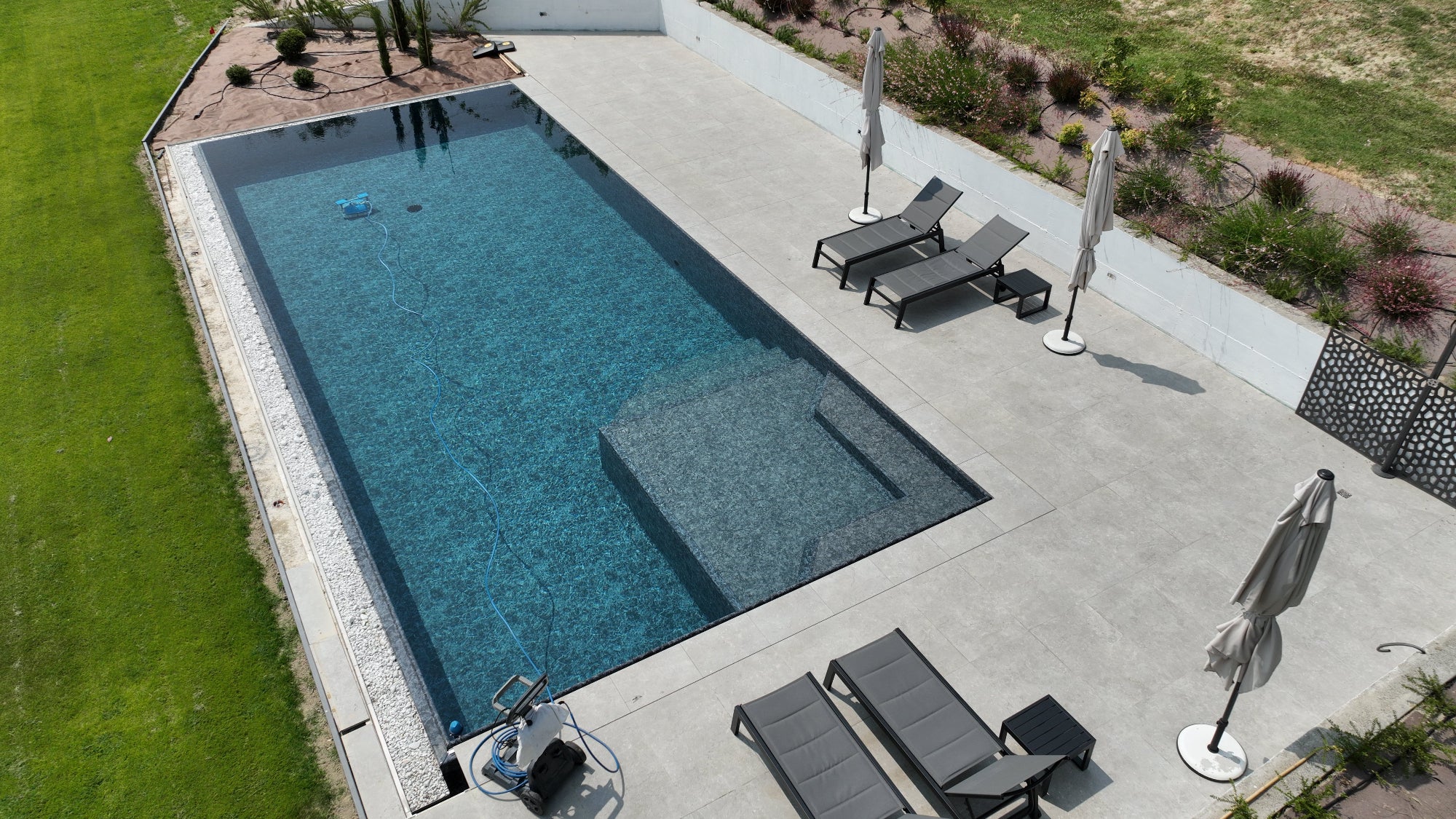 piscina a sfioro senza vasca di compenso dal design minimal e moderno. Vaschette con sassi decorativi su due lati e rivestimento nero marmorizzato. Spiaggia interna in angolo.