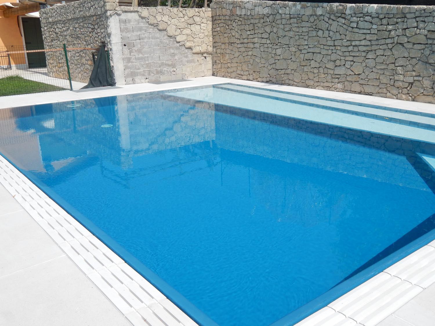 Piscina a sfioro isotermica con rivestimento Sea Blu e scalini longitudinali chiari. La piscina è impreziosita da un bordo in pietra ricostruita che riprende le tonalità chiare della pavimentazione circostante.