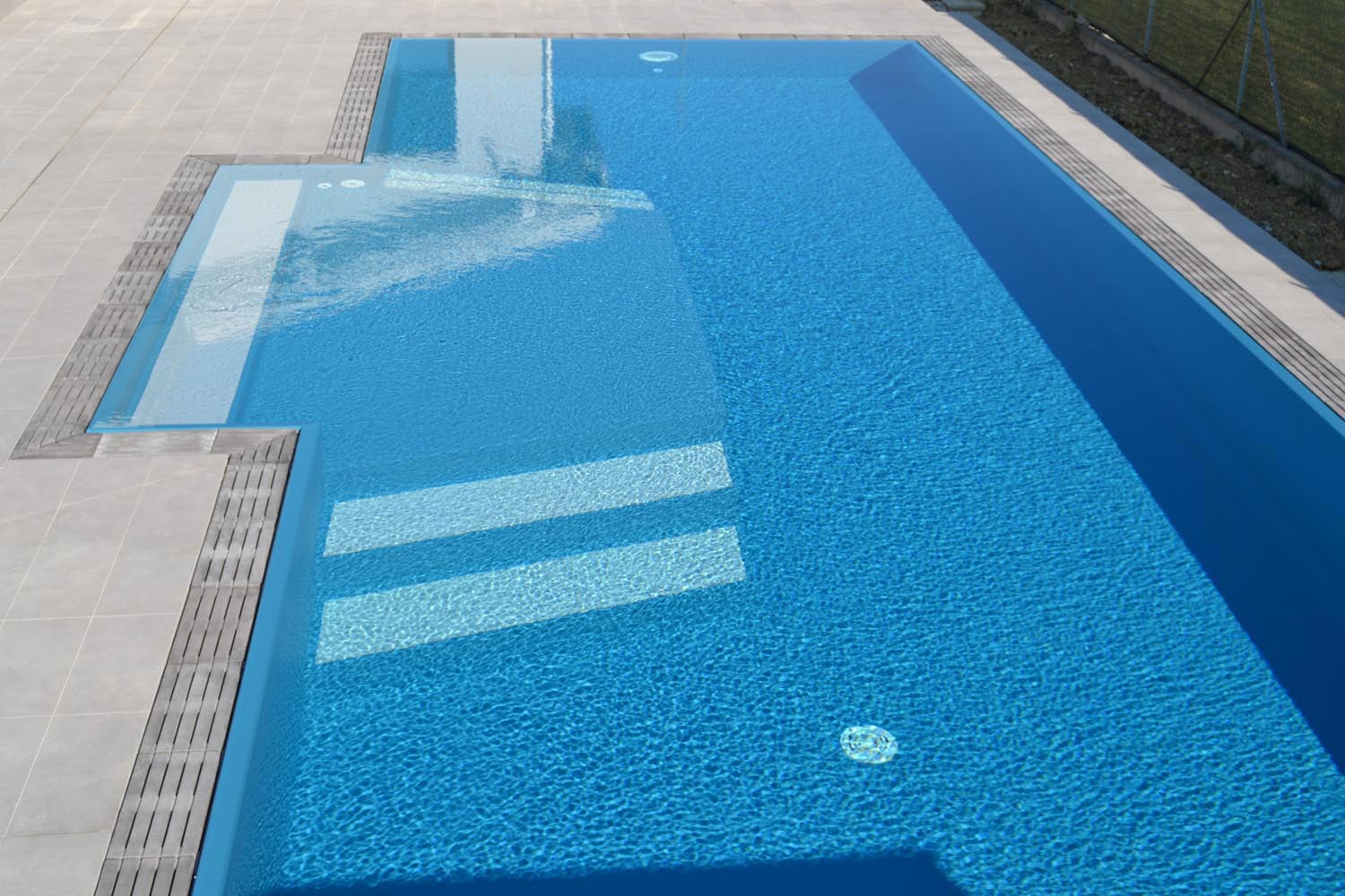 Dettaglio bagnasciuga interno con scalini esterni bianchi. La forma permette di rilassarsi con sdraio o lettino e scendere in piscina in tutta sicurezza grazie al rivestimento antiscivolo.