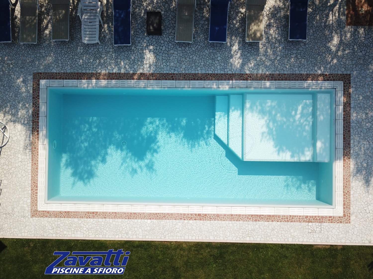 Vista dall'alto di una piscina a sfioro Zavatti con griglia in pietra ricostruita bianca e zona relax interna