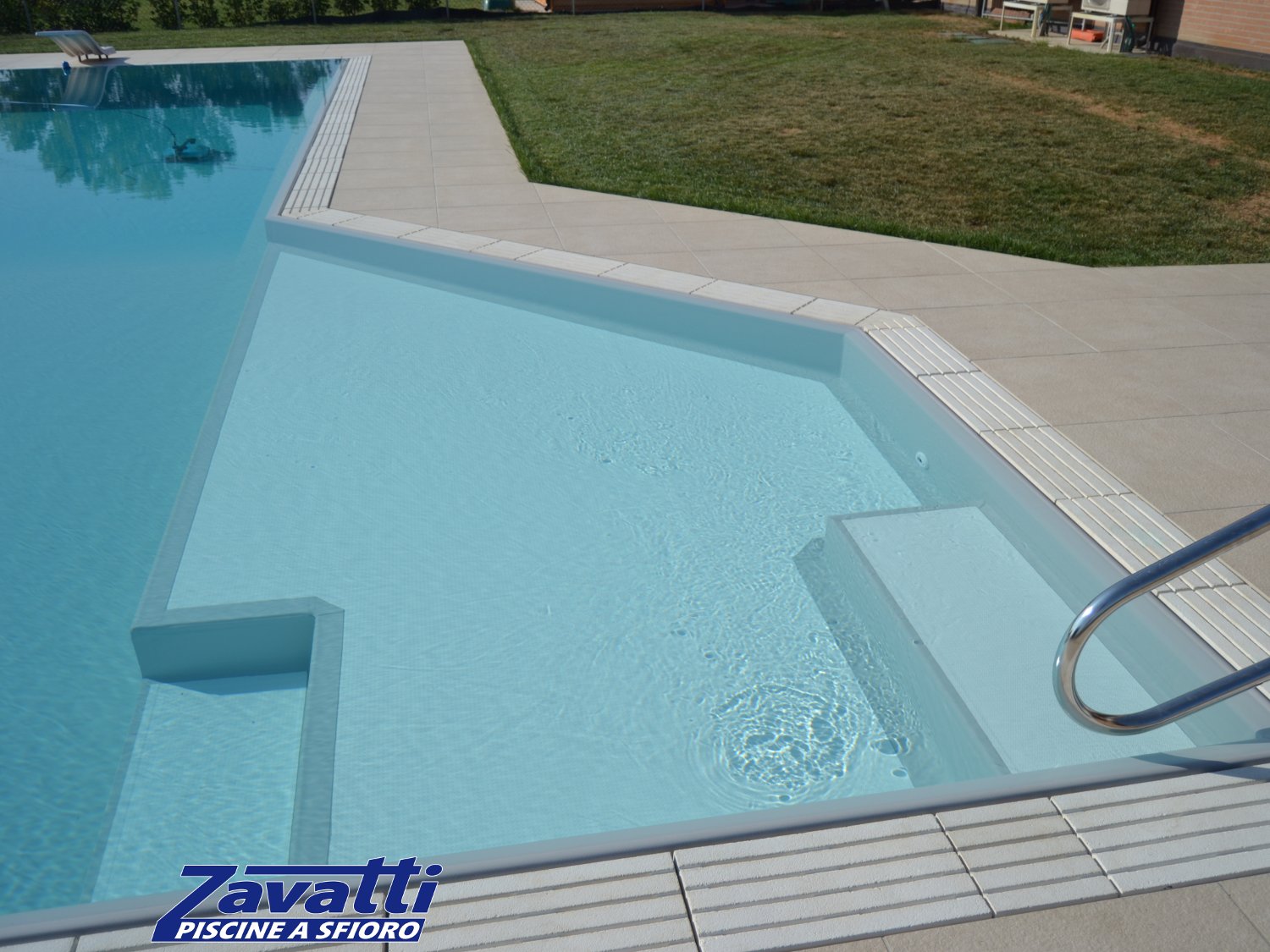 Zona relax piscina a sfioro realizzata con liner grigio. Particolare design dovuto alla forma della piscina e agli scalini bianchi